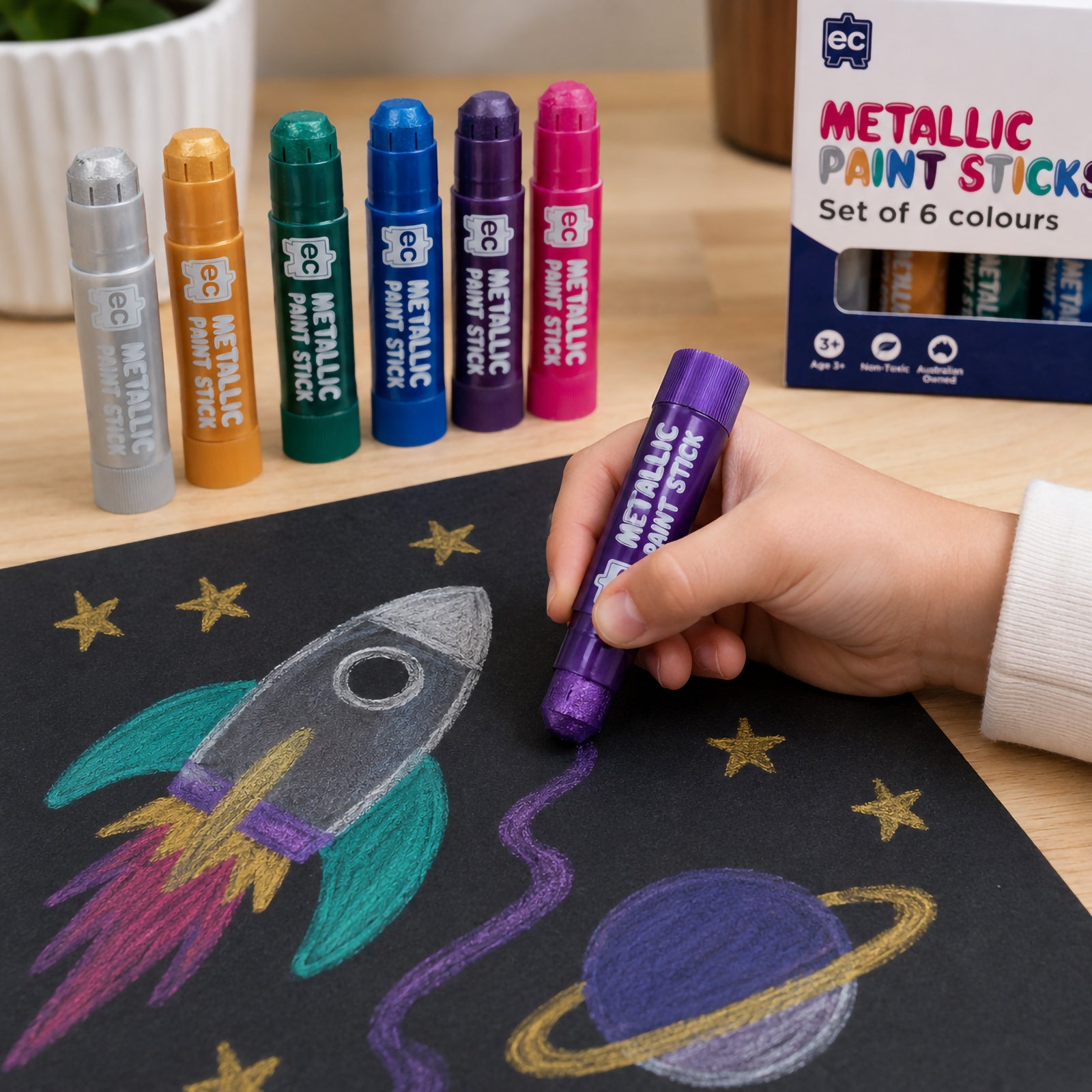 Child drawing a metallic space scene with rocket and planets on black paper using purple metallic paint stick, with full paint stick set and packaging in background.