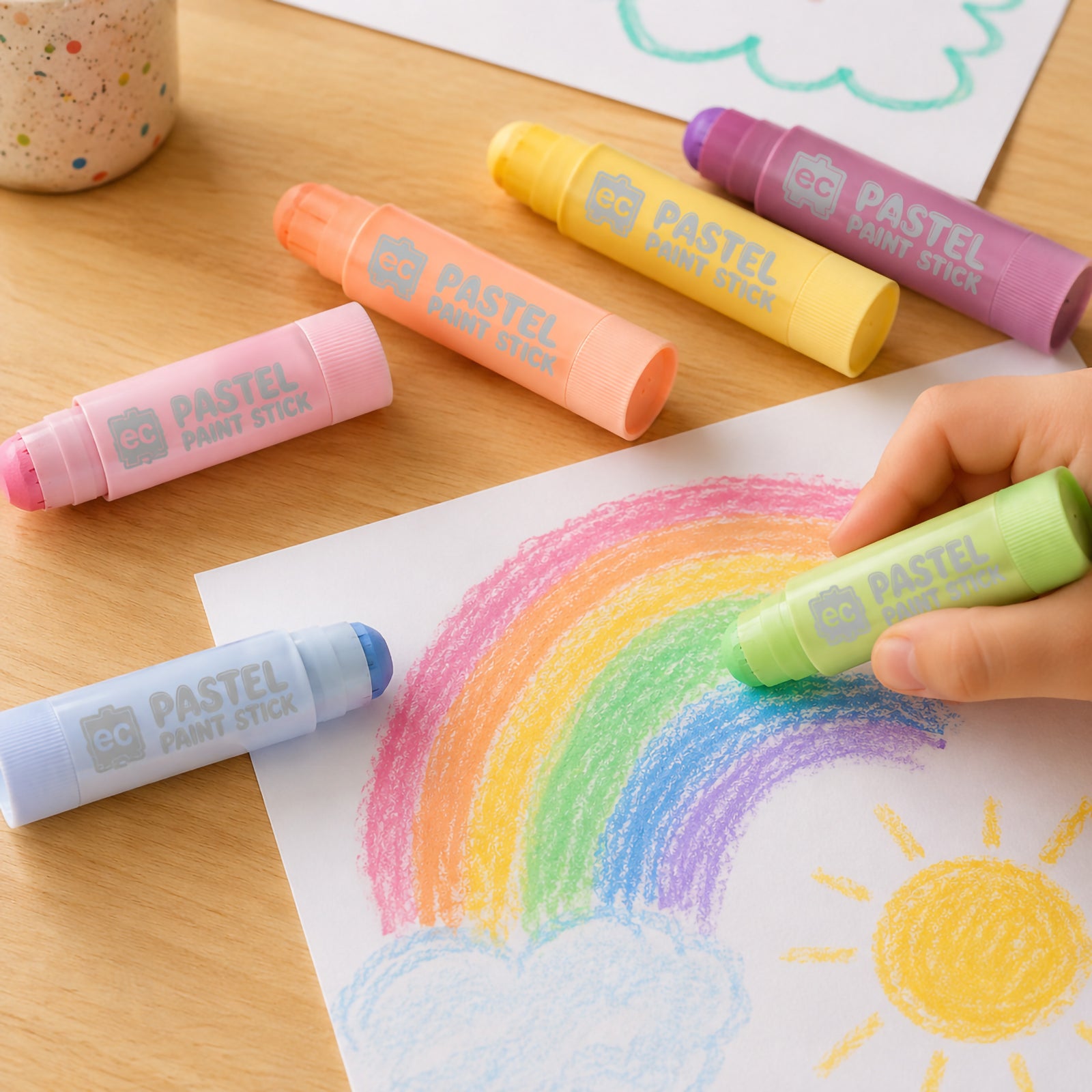 Child using a green pastel paint stick to colour a rainbow drawing on paper. Several pastel paint sticks are scattered across a wooden table with playful artwork including sunshine and clouds in soft pastel tones.
