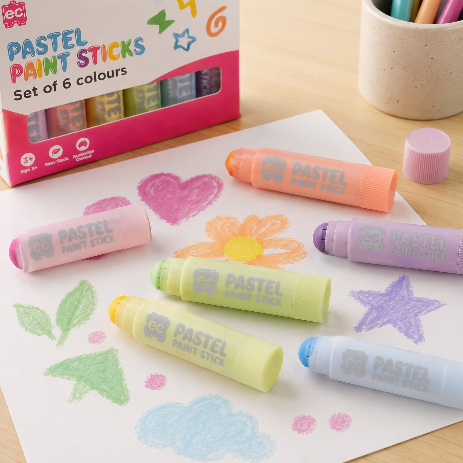 Lifestyle image showing Educational Colours Pastel Paint Sticks arranged on a sheet of paper with pastel drawings including hearts, flowers, stars and clouds. The soft coloured paint sticks are displayed beside the product packaging on a wooden desk during a creative art activity.