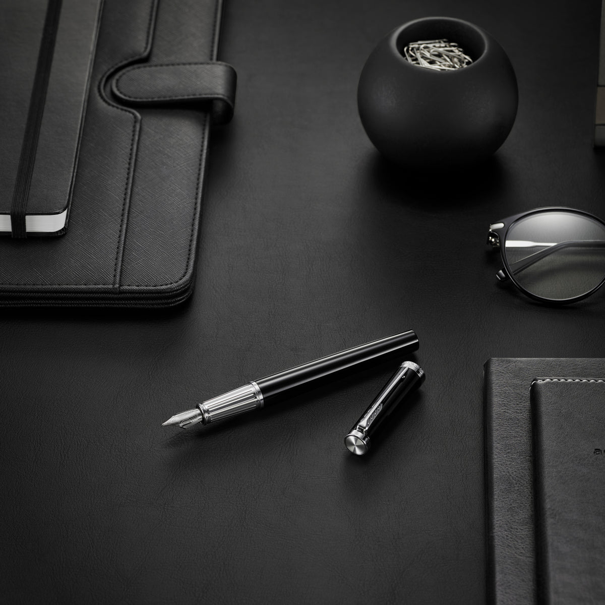 Black and chrome Ingenuity fountain pen and cap lying on a dark desk beside a notebook and glasses