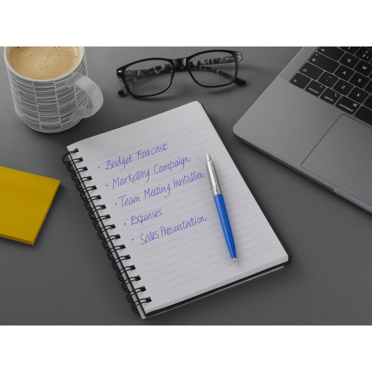 Blue Parker Jotter on a spiral notebook with a list in blue ink, coffee mug, glasses and laptop on a grey desk