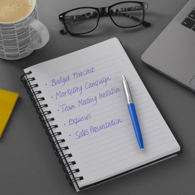 Blue Parker Jotter on a spiral notebook with a blue handwritten list, glasses and coffee cup behind