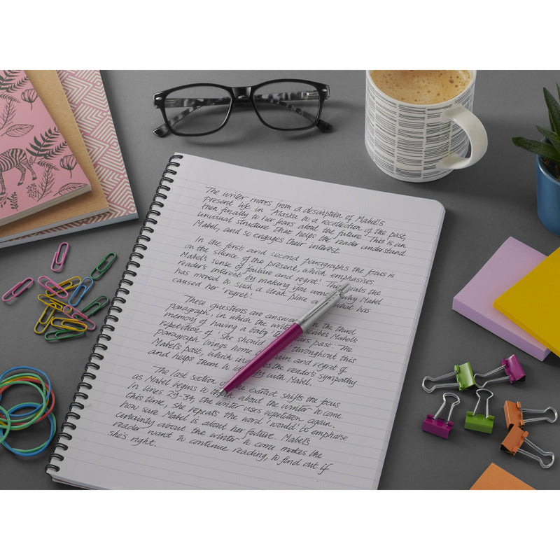 Pink Parker Jotter on a spiral notebook of black handwriting, with glasses, mug, clips and sticky notes on a grey desk