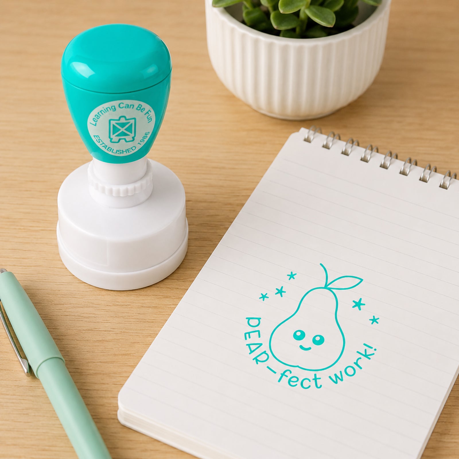 Learning Can Be Fun Pear-fect Work Merit Stamp displayed beside a notepad and pastel pen on a classroom desk. The teal self inking stamp creates a cheerful pear themed reward impression designed to celebrate excellent student work and encourage positive learning outcomes.