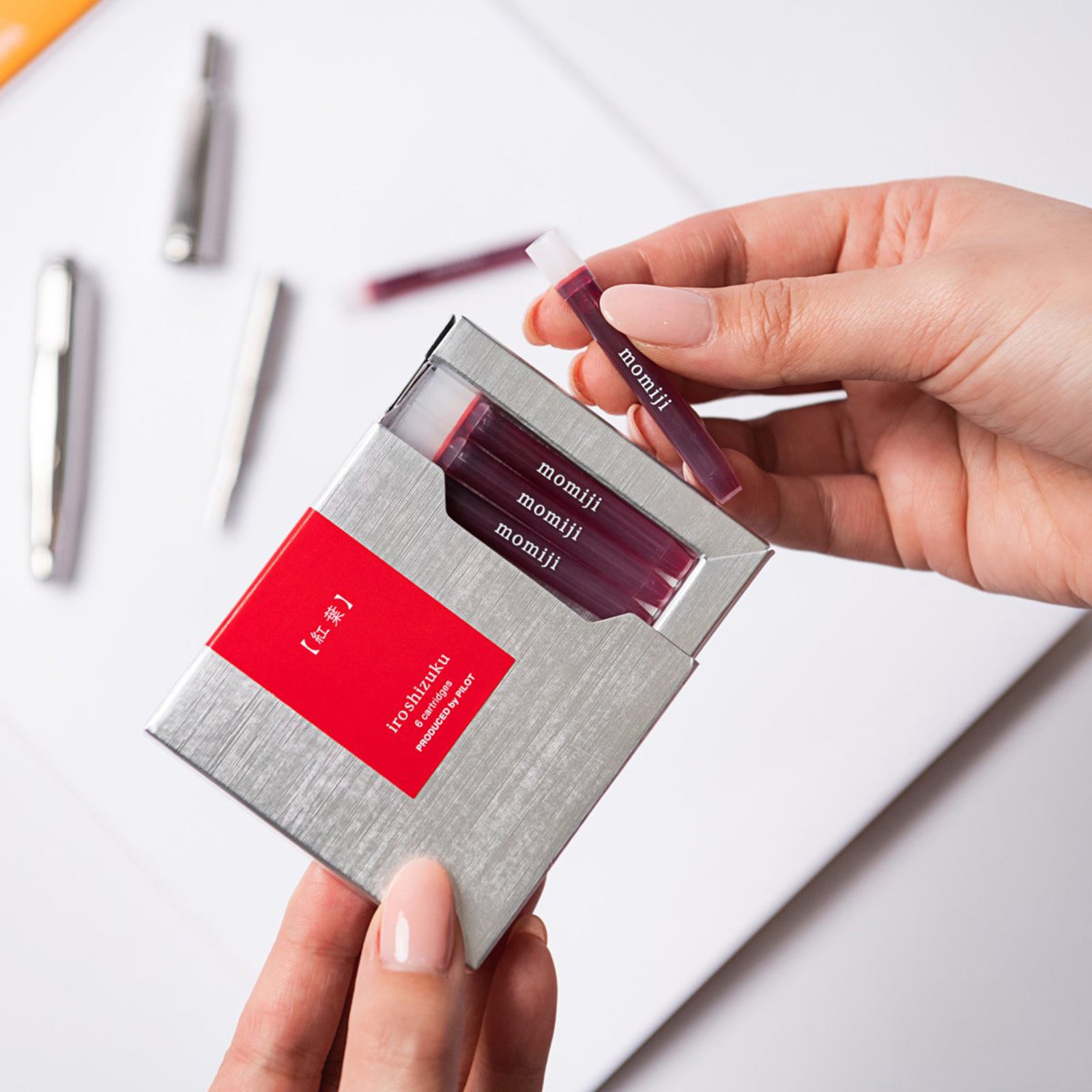 A hand removes a Pilot Iroshizuku Momiji fountain pen ink cartridge from the premium six cartridge presentation box, demonstrating the convenient proprietary cartridge design for quick, clean fountain pen refilling.
