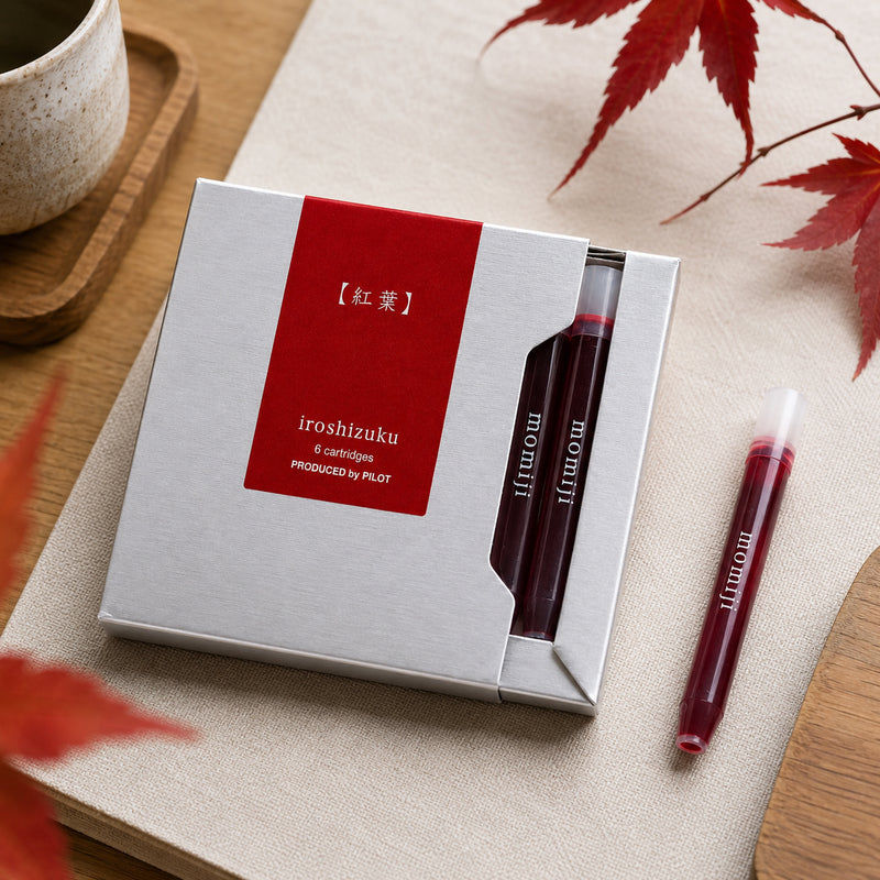 Close up lifestyle image of the Pilot Iroshizuku Momiji six cartridge presentation box resting on a neutral linen surface with autumn maple leaves and a ceramic cup. One crimson ink cartridge is positioned beside the premium packaging to highlight the elegant seasonal colour.