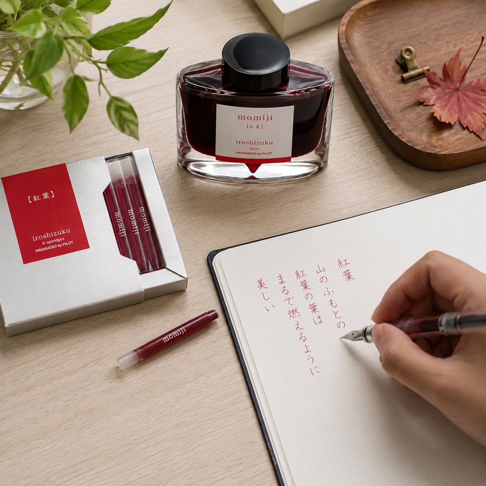 Pilot Iroshizuku Momiji fountain pen ink cartridges displayed alongside the matching 50ml ink bottle on a timber writing desk while a writer uses a compatible fountain pen to create elegant Japanese calligraphy. The premium cartridge box and deep crimson autumn inspired ink showcase the refined craftsmanship of the Iroshizuku collection.