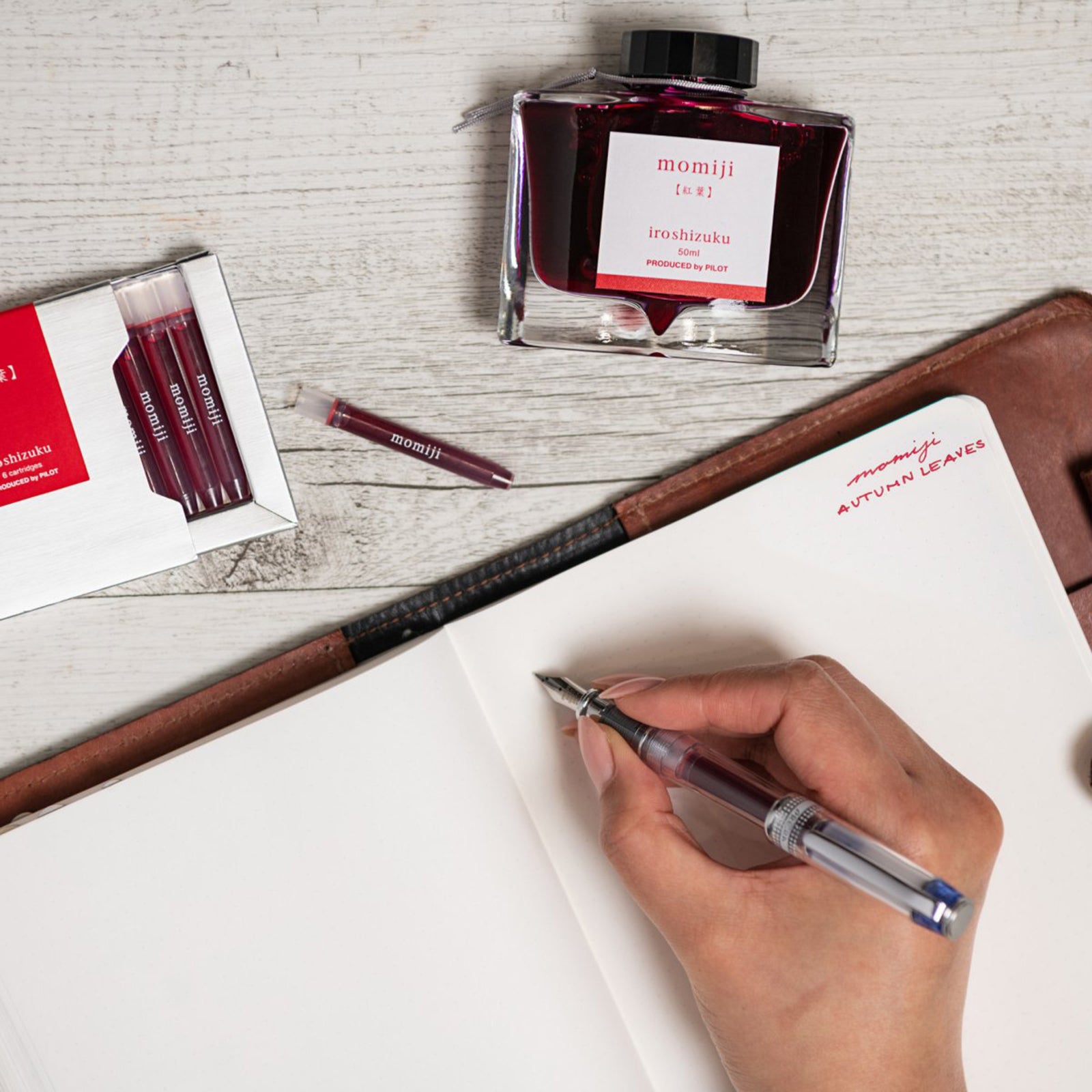 Creative writing workspace featuring the Pilot Iroshizuku Momiji cartridge box, matching 50ml ink bottle, loose cartridge and compatible fountain pen beside an open notebook. The warm autumn inspired scene highlights the rich crimson ink for journalling and creative writing.