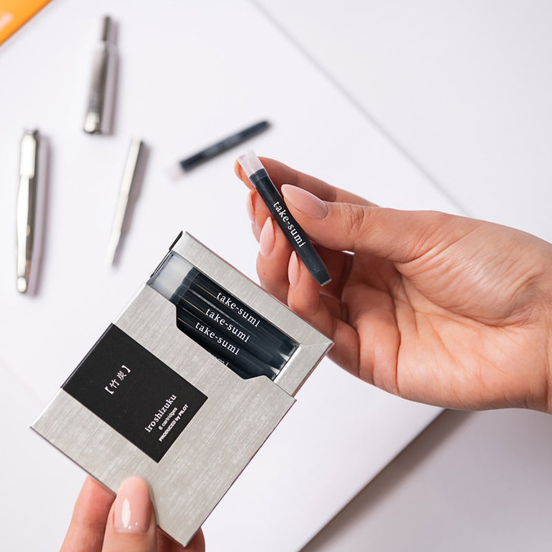 A hand removes a Pilot Iroshizuku Take Sumi fountain pen ink cartridge from the premium six cartridge presentation box, demonstrating the convenient proprietary cartridge design for quick and clean fountain pen refilling.