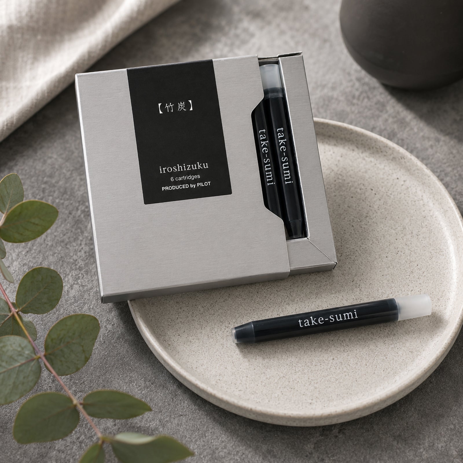Lifestyle close up of the Pilot Iroshizuku Take Sumi six cartridge presentation box resting on a ceramic plate with a single black ink cartridge displayed beside it. Soft neutral styling and natural greenery emphasise the premium Japanese design.