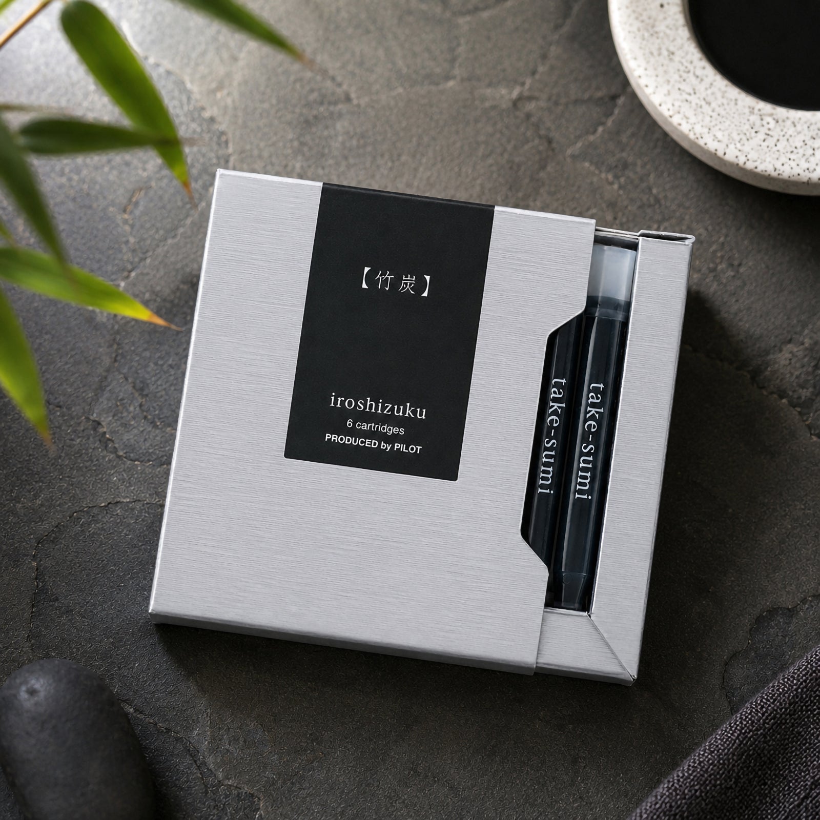 Top view of the Pilot Iroshizuku Take Sumi fountain pen cartridge gift box placed on a dark stone surface with bamboo leaves, smooth black stones and ceramic tableware. The premium packaging and visible cartridges create a refined Japanese inspired display.
