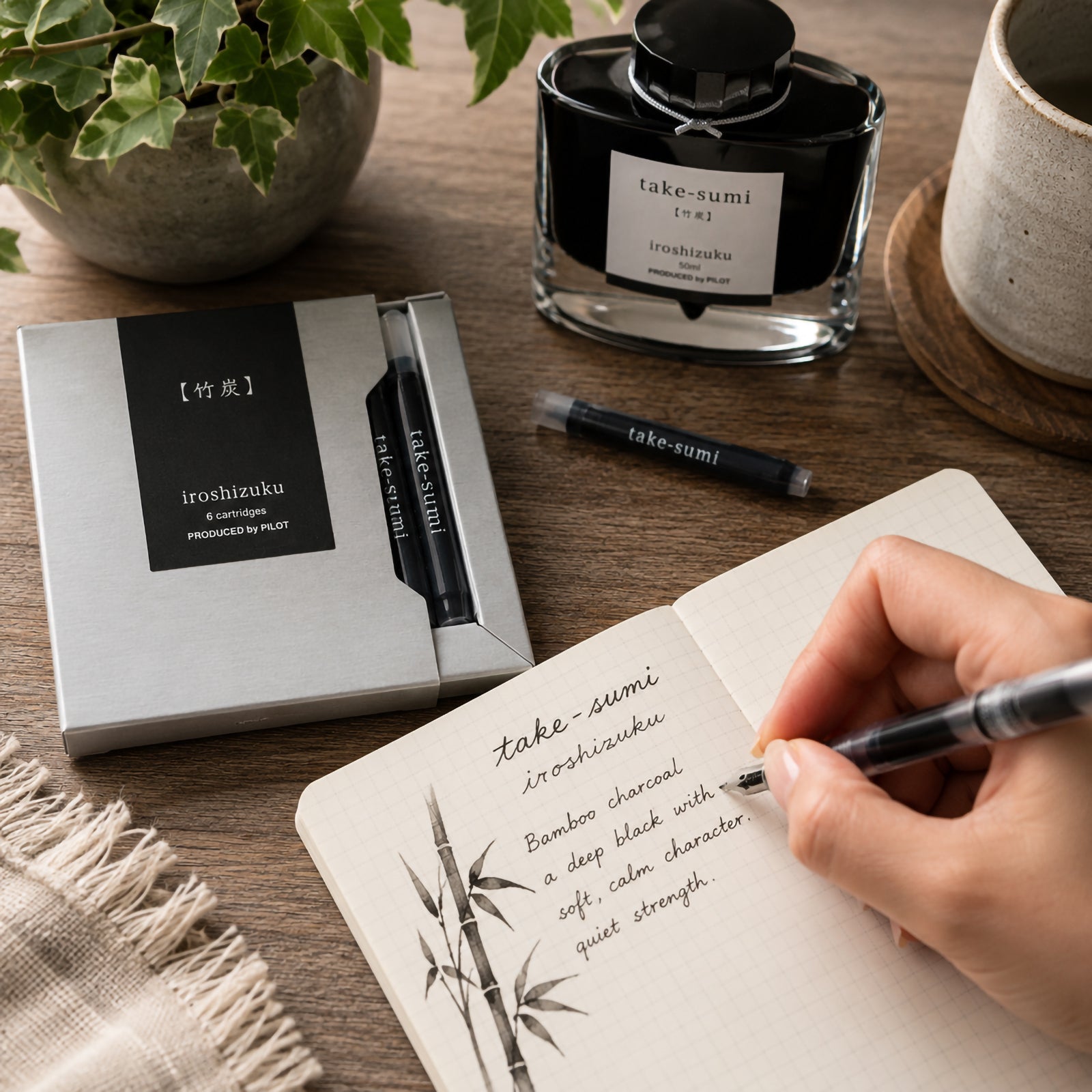 Pilot Iroshizuku Take Sumi fountain pen ink cartridges displayed beside the matching 50ml ink bottle on a wooden writing desk while a hand writes in a notebook with a compatible fountain pen. The elegant bamboo themed workspace highlights the deep black ink inspired by Japanese bamboo charcoal.