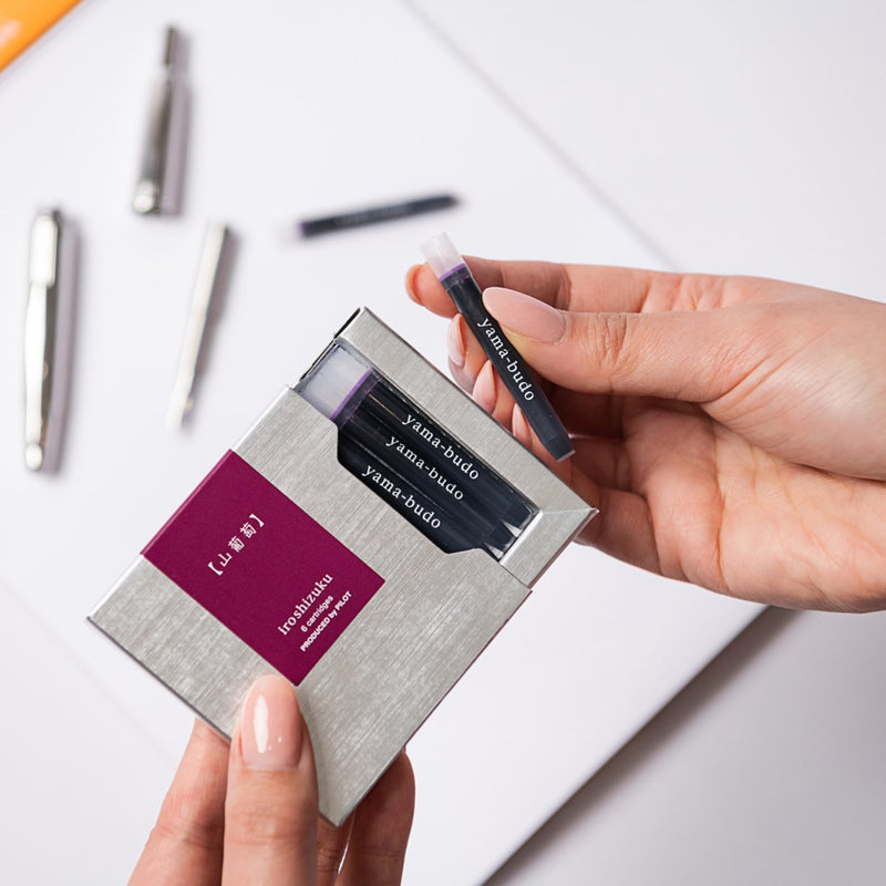 A hand removes a Pilot Iroshizuku Yama Budo fountain pen ink cartridge from the premium six cartridge presentation box, demonstrating the convenient proprietary cartridge design for quick and clean fountain pen refilling.