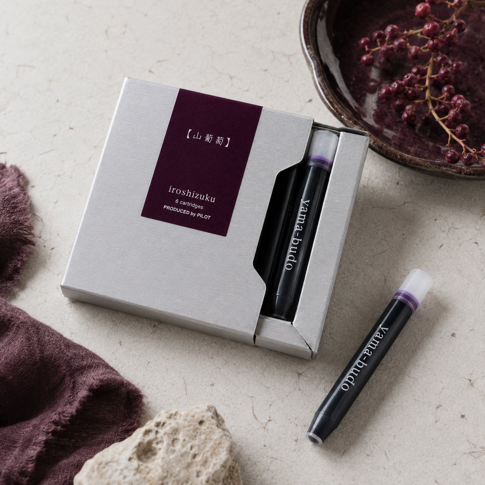 Lifestyle close up of the Pilot Iroshizuku Yama Budo six cartridge presentation box resting on a natural stone surface beside a ceramic bowl filled with dark grapes. A single purple crimson ink cartridge highlights the premium Japanese packaging and distinctive colour.
