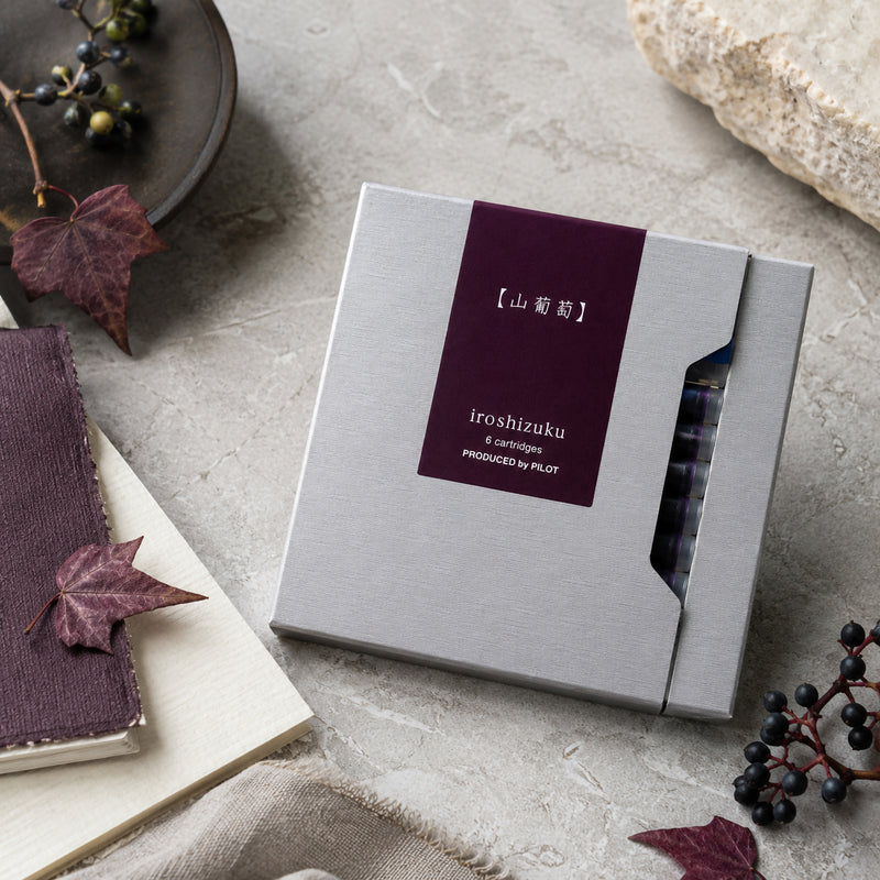 Top view of the Pilot Iroshizuku Yama Budo fountain pen cartridge gift box arranged on a textured stone surface with vine leaves, berries and natural styling elements. The premium packaging reflects the inspiration of the Crimson Glory Vine colour.