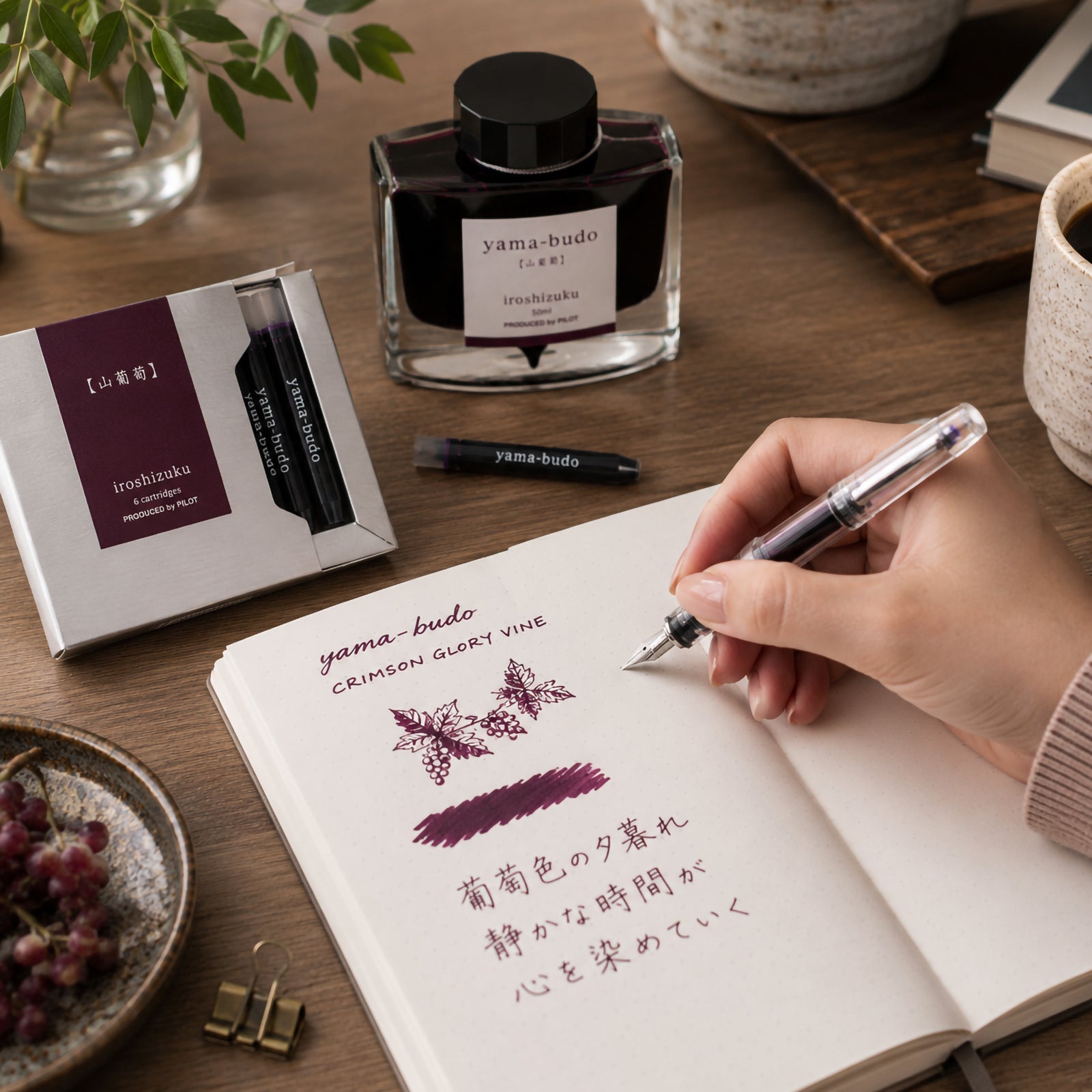 Pilot Iroshizuku Yama Budo fountain pen ink cartridges displayed beside the matching 50ml ink bottle on a wooden writing desk while a hand writes in a notebook using a compatible fountain pen. Rich purple crimson ink inspired by the Japanese Crimson Glory Vine is showcased in an elegant creative workspace.
