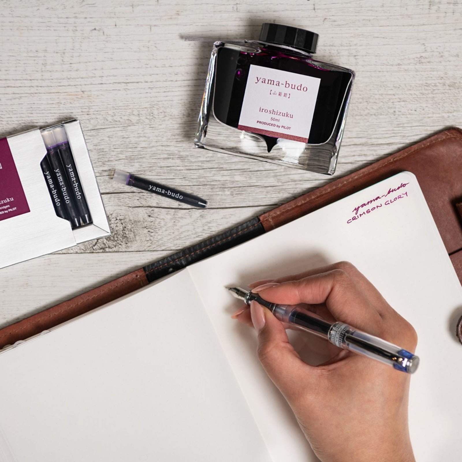 Creative writing workspace featuring the Pilot Iroshizuku Yama Budo cartridge box, matching 50ml ink bottle, loose cartridge and compatible fountain pen beside an open notebook. The scene celebrates expressive writing with rich purple crimson ink inspired by Japanese nature.