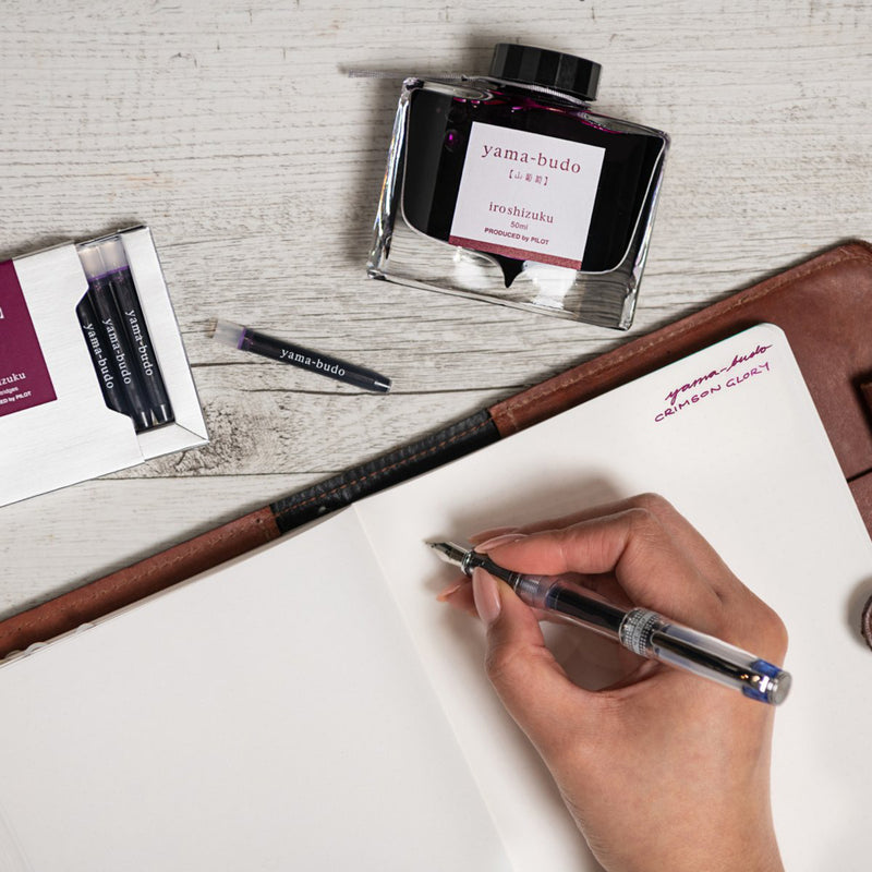 Creative writing workspace featuring the Pilot Iroshizuku Yama Budo cartridge box, matching 50ml ink bottle, loose cartridge and compatible fountain pen beside an open notebook. The scene celebrates expressive writing with rich purple crimson ink inspired by Japanese nature.