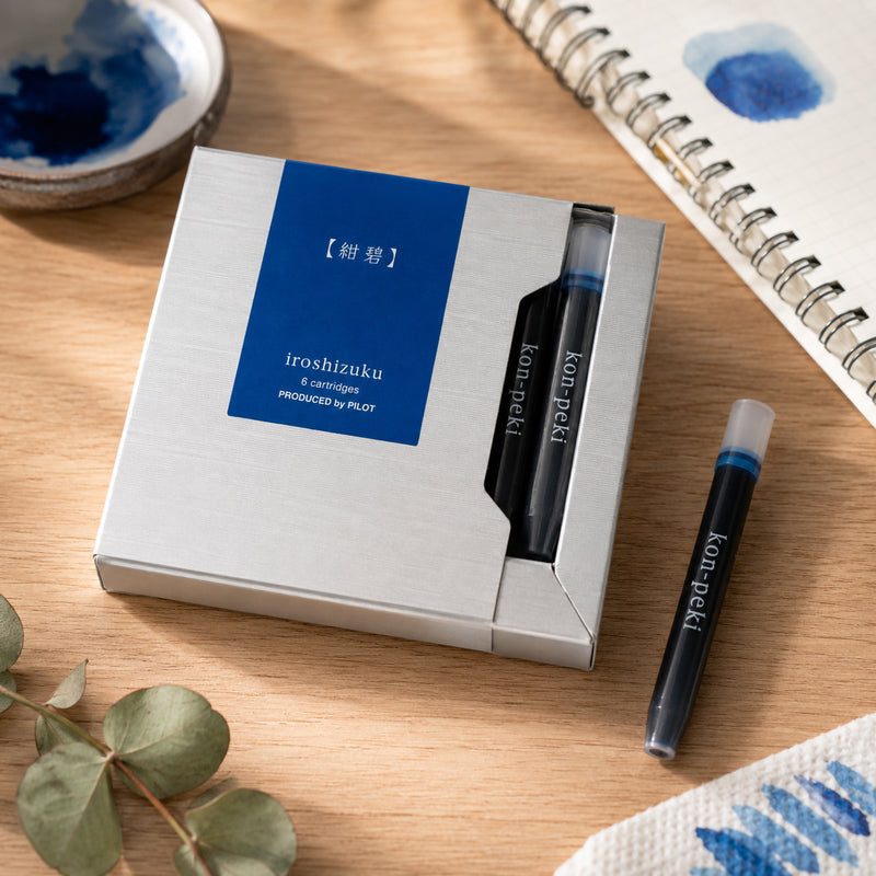Lifestyle close up of the Pilot Iroshizuku Kon Peki six cartridge presentation box resting on a wooden desktop beside a sketchbook with blue ink swatches. A single deep cerulean blue cartridge is displayed to showcase the premium Japanese packaging and brilliant ink colour.