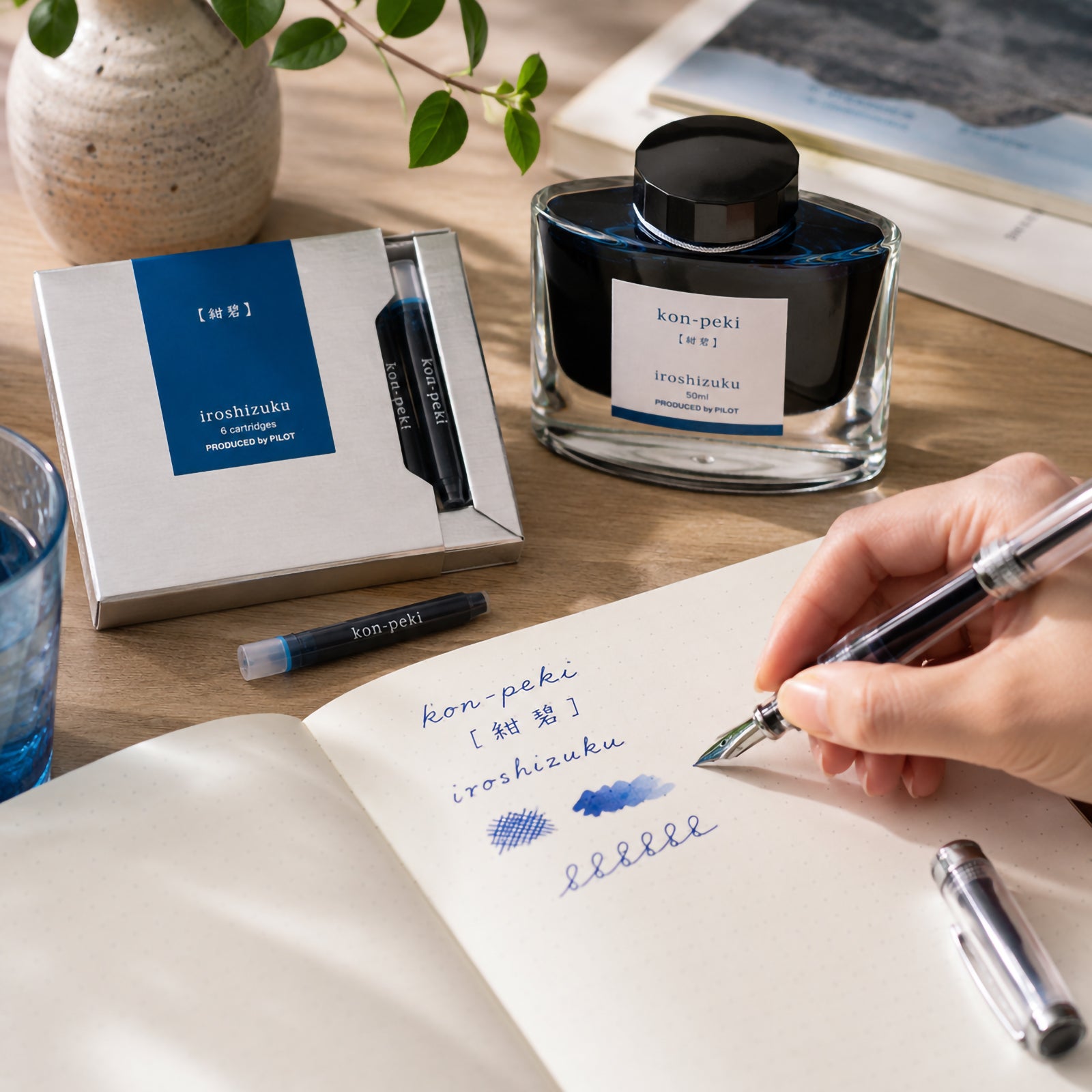 Pilot Iroshizuku Kon Peki fountain pen ink cartridges displayed beside the matching 50ml ink bottle on a bright wooden writing desk while a hand writes with a compatible fountain pen. The vivid cerulean blue ink samples highlight the fresh, vibrant colour inspired by clear Japanese skies.