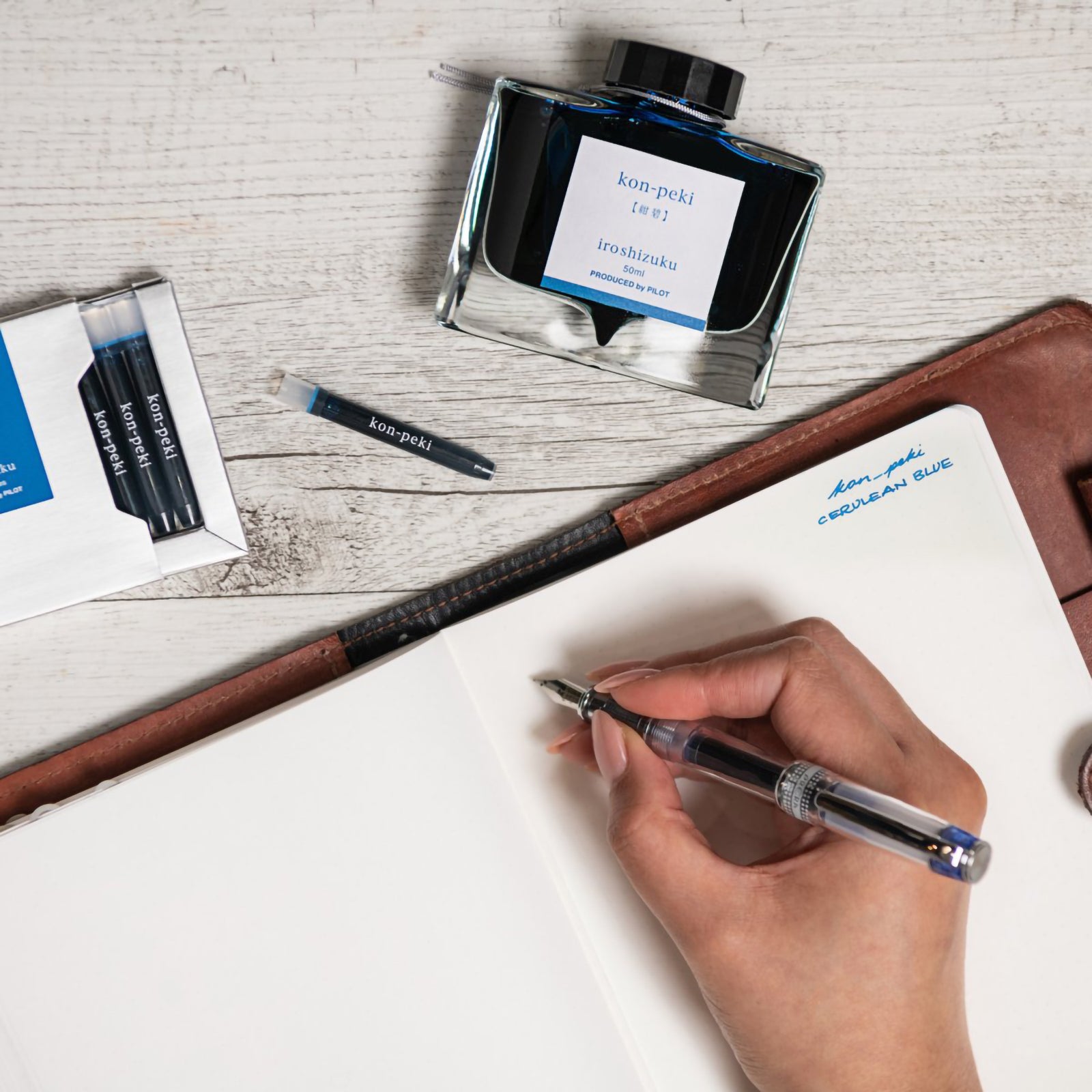 Creative writing workspace featuring the Pilot Iroshizuku Kon Peki cartridge box, matching 50ml ink bottle, loose cartridge and compatible fountain pen beside an open notebook. The scene celebrates expressive writing using rich cerulean blue ink inspired by the beauty of clear skies.
