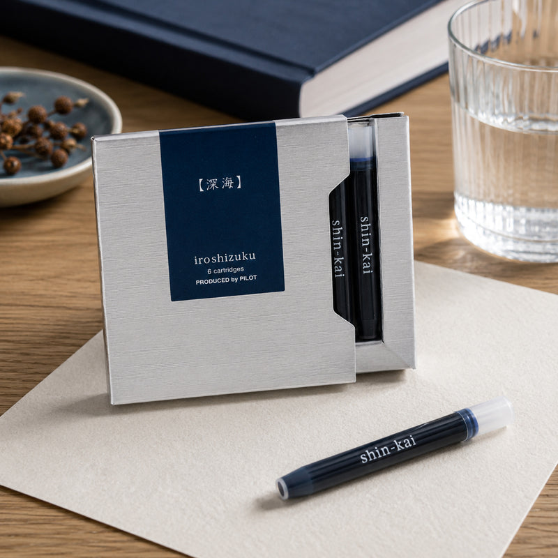 Lifestyle close up of the Pilot Iroshizuku Shin Kai six cartridge presentation box resting on a desk beside a notebook and a single blue black cartridge. The premium packaging highlights the refined Deep Sea colour and genuine Japanese craftsmanship.