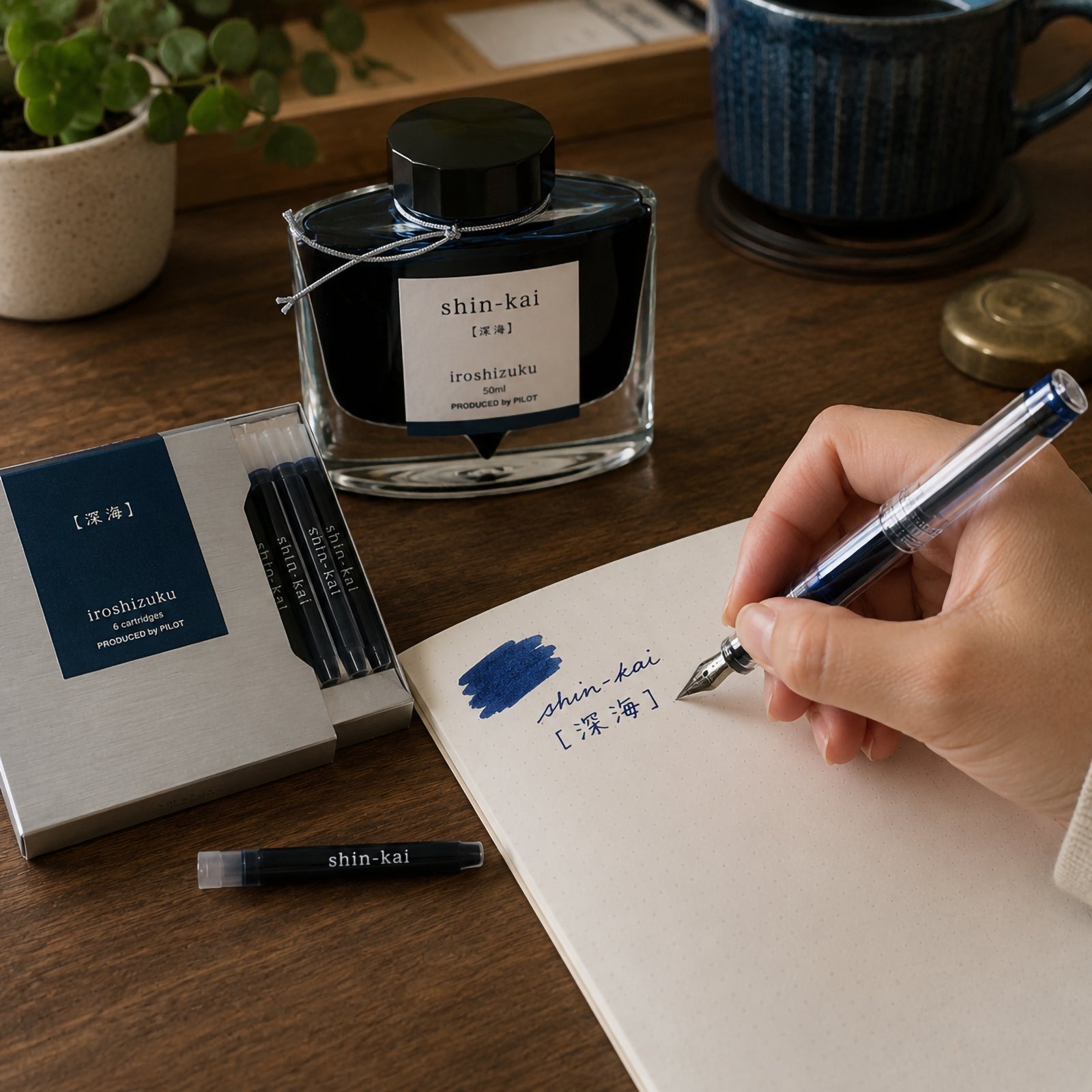 Pilot Iroshizuku Shin Kai fountain pen ink cartridges displayed beside the matching 50ml ink bottle on a wooden writing desk while a hand writes with a compatible fountain pen. Rich blue black ink samples demonstrate the elegant Deep Sea colour inspired by the mysterious Japanese ocean.
