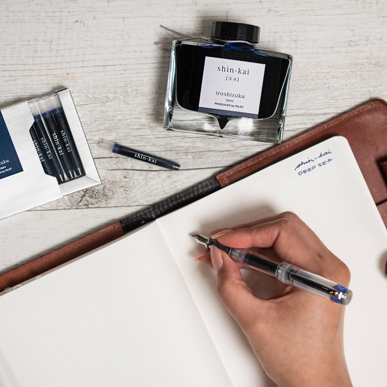 Creative writing workspace featuring the Pilot Iroshizuku Shin Kai cartridge box, matching 50ml ink bottle, loose cartridge and compatible fountain pen beside an open notebook. The scene showcases expressive writing with the rich Deep Sea blue black ink.