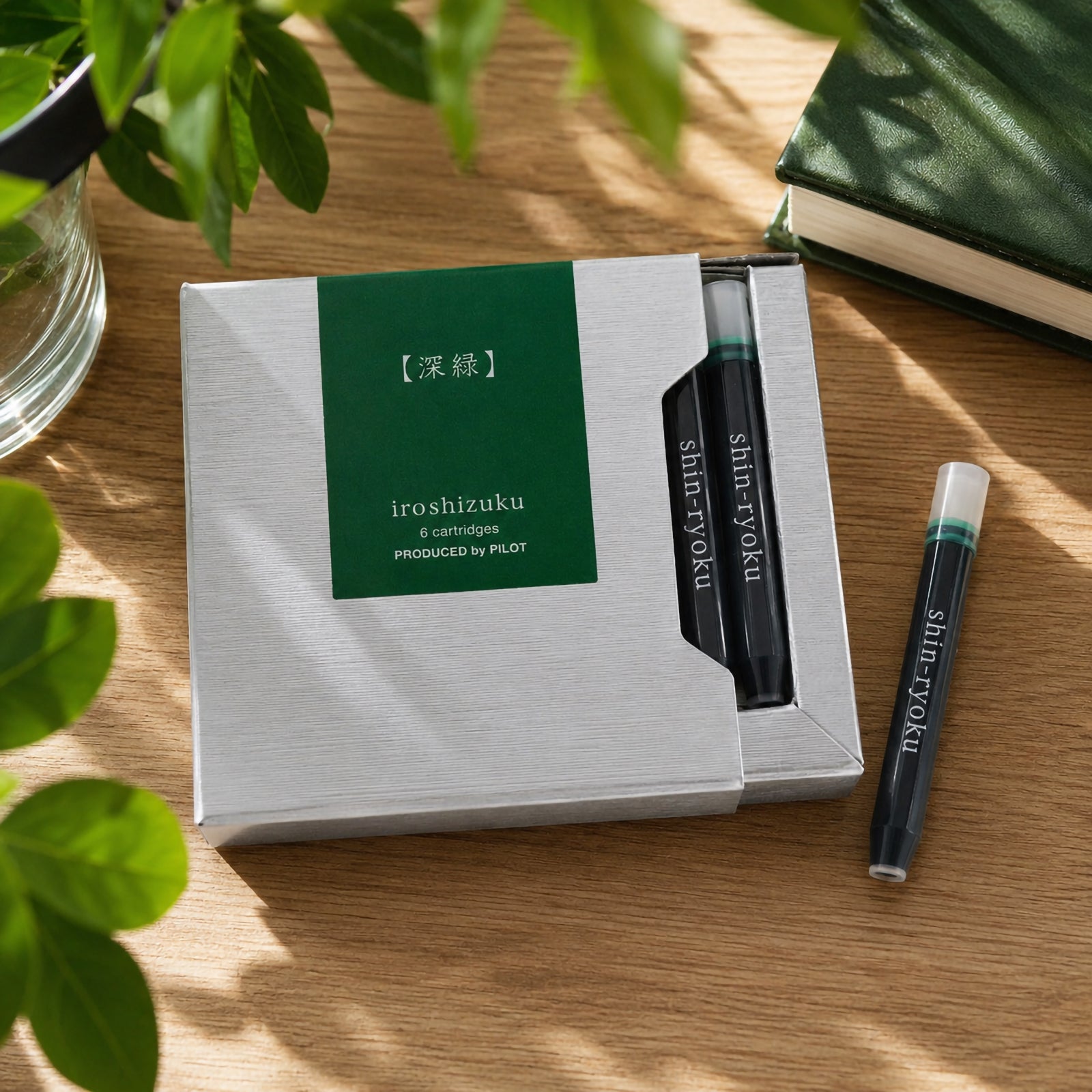 Lifestyle close up of the Pilot Iroshizuku Shin Ryoku six cartridge presentation box resting on a timber desk surrounded by green foliage, a notebook and a single forest green cartridge. The premium packaging highlights the elegant Japanese design.