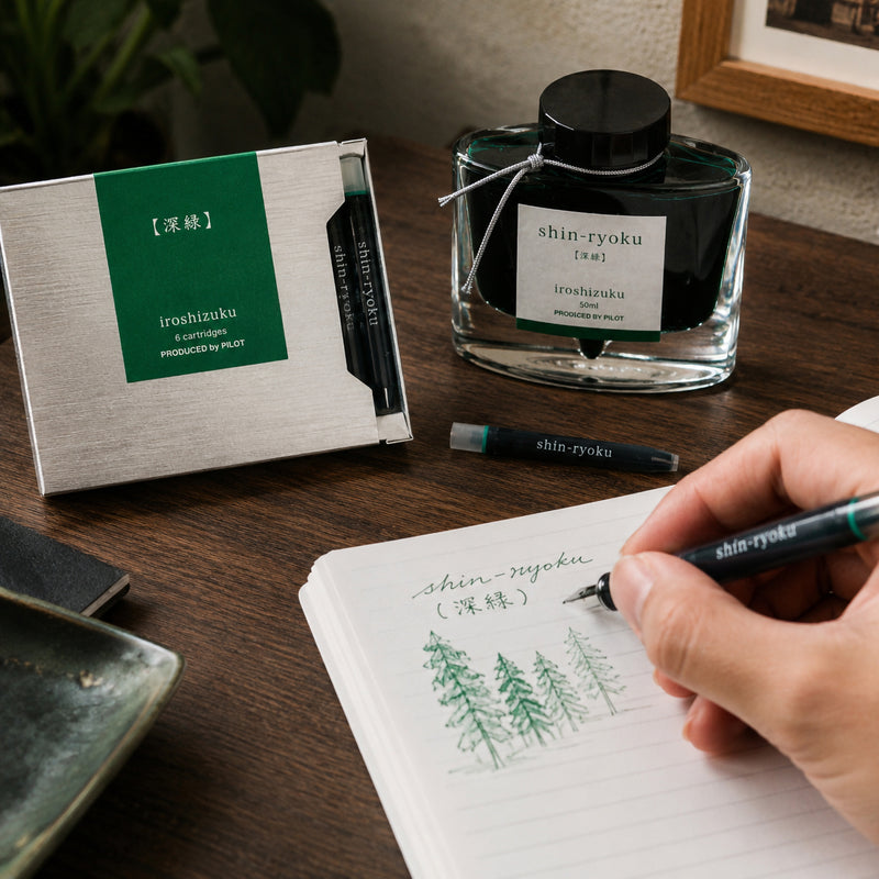 Pilot Iroshizuku Shin Ryoku fountain pen ink cartridges displayed beside the matching 50ml ink bottle on a wooden writing desk while a hand sketches evergreen trees with a compatible fountain pen. The rich forest green ink demonstrates smooth flow and nature inspired colour.