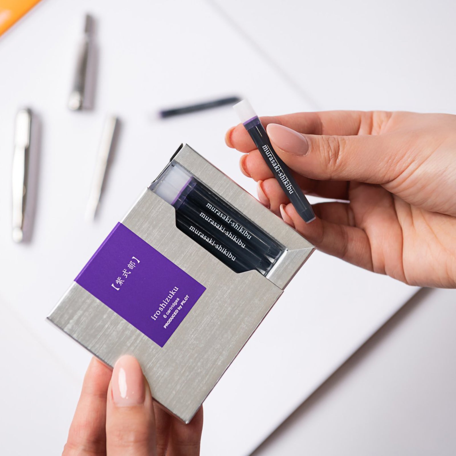 A hand removes a genuine Pilot Iroshizuku Murasaki Shikibu fountain pen ink cartridge from the premium six cartridge presentation box, demonstrating the convenient proprietary cartridge system for quick and clean fountain pen refilling.