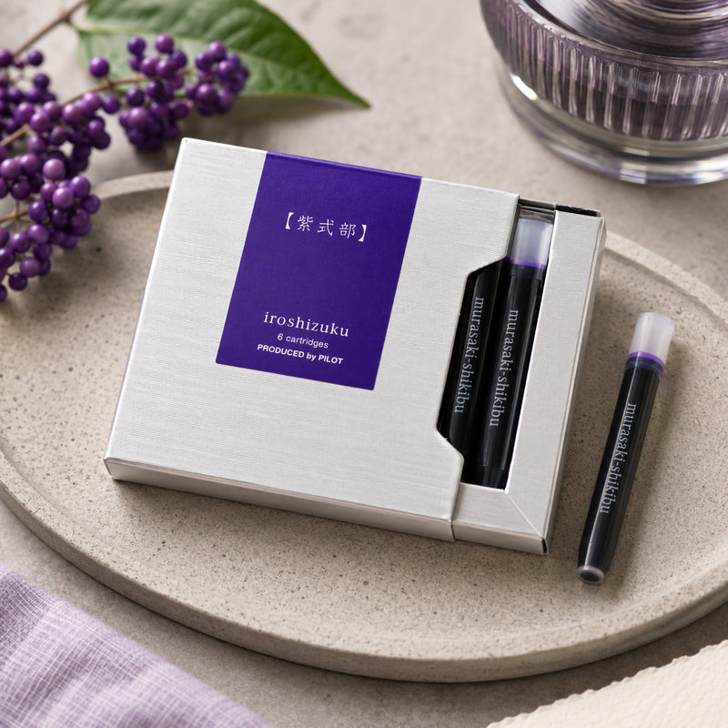 Premium lifestyle display of the Pilot Iroshizuku Murasaki Shikibu six cartridge presentation box resting on a ceramic tray with Japanese beautyberry branches and a single purple ink cartridge. The scene highlights the elegant packaging and sophisticated colour inspiration.