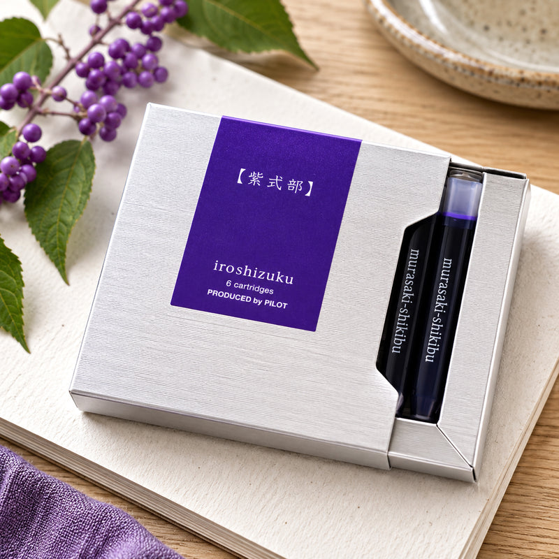 Pilot Iroshizuku Murasaki Shikibu fountain pen ink cartridge presentation box displayed on an open notebook with decorative beautyberry branches and soft natural lighting. The premium packaging reflects the graceful Japanese inspired design.