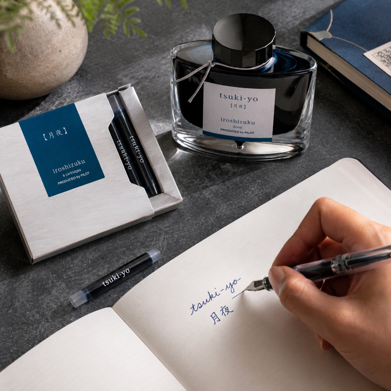 Pilot Iroshizuku Tsuki yo Moonlight fountain pen ink cartridges displayed beside the matching 50ml ink bottle while a hand writes with a compatible fountain pen in a notebook. The elegant blue black ink reflects the calm atmosphere of a moonlit evening.