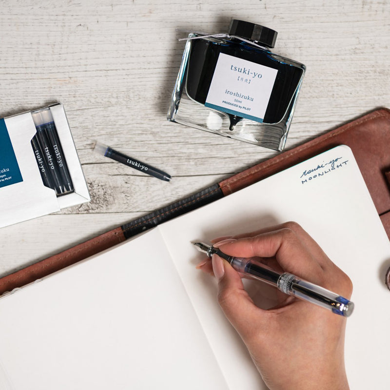 Creative workspace featuring the Pilot Iroshizuku Tsuki yo cartridge box, matching 50ml ink bottle, loose cartridge and compatible fountain pen beside an open notebook. The arrangement showcases the rich Moonlight blue black ink for elegant writing and creative projects.