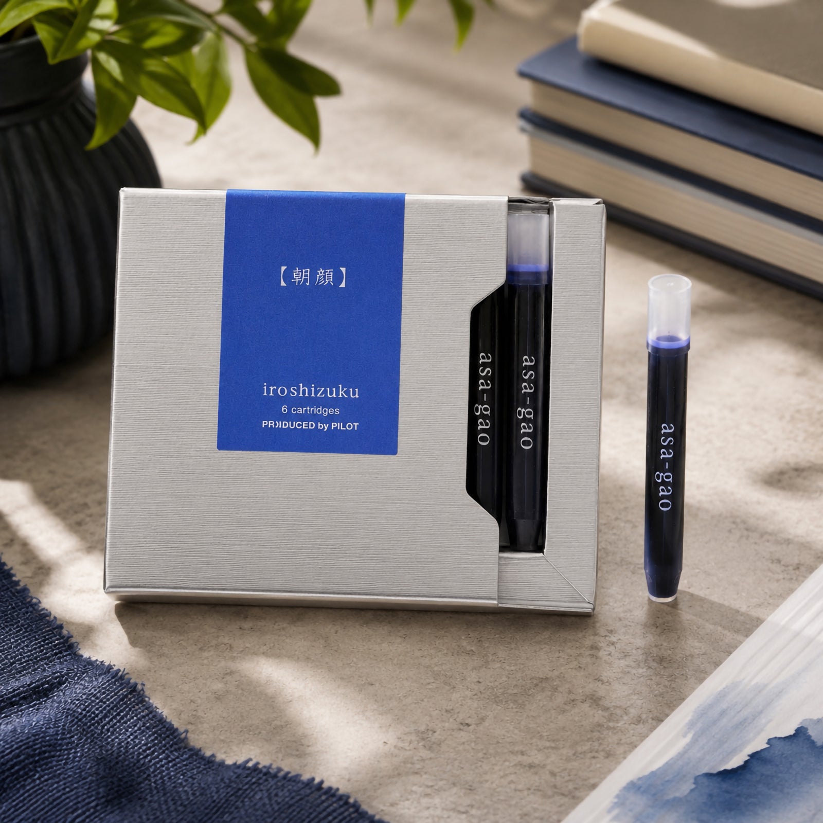Premium lifestyle presentation of the Pilot Iroshizuku Asa gao six cartridge gift box with two visible cartridges and one loose cartridge beside stacked notebooks and botanical styling. The scene highlights the elegant packaging and rich blue ink colour.