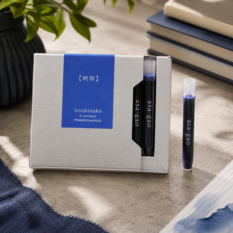 Premium lifestyle presentation of the Pilot Iroshizuku Asa gao six cartridge gift box with two visible cartridges and one loose cartridge beside stacked notebooks and botanical styling. The scene highlights the elegant packaging and rich blue ink colour.