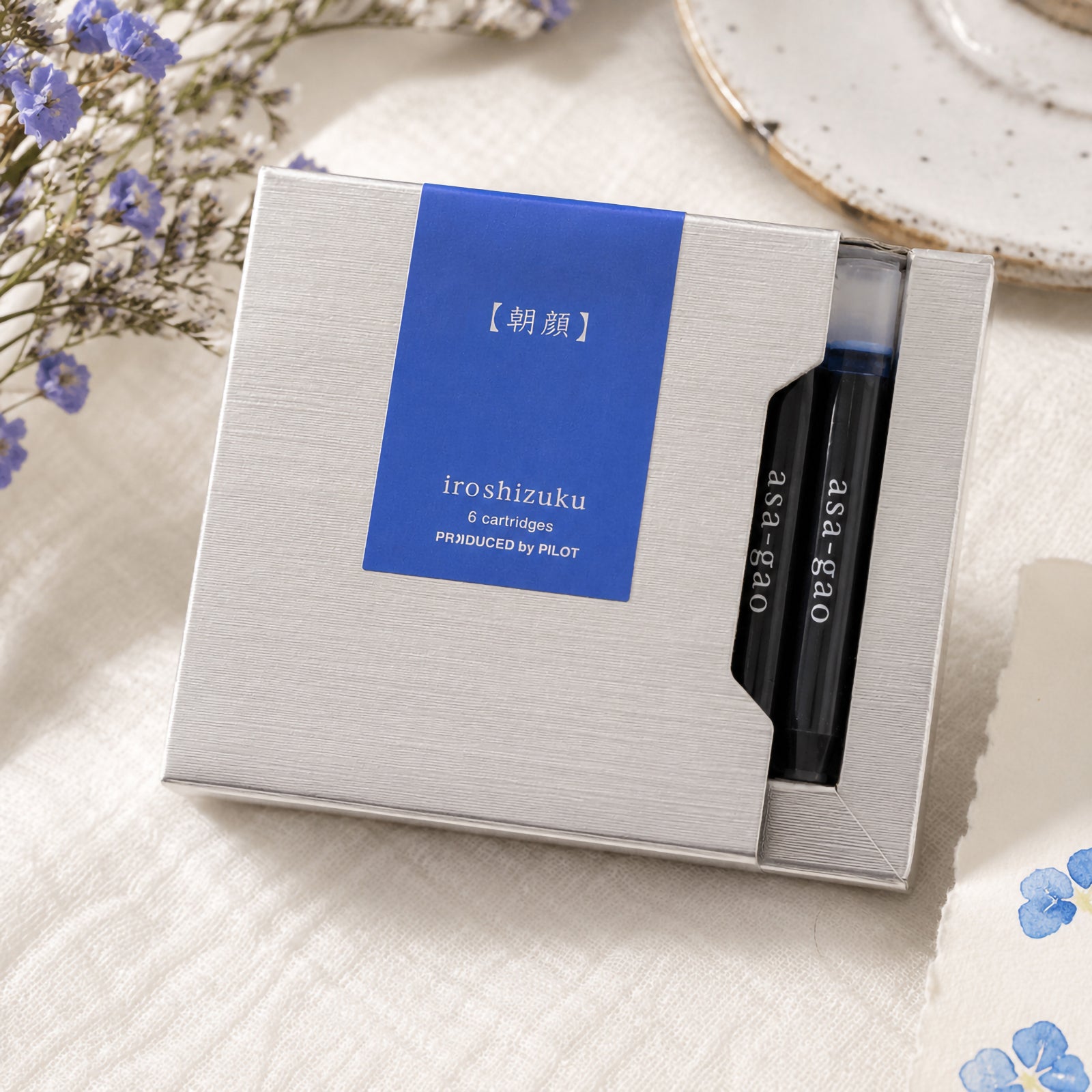 Pilot Iroshizuku Asa gao fountain pen cartridge presentation box displayed on a light fabric surface surrounded by delicate blue flowers and stationery. The composition reinforces the Morning Glory inspiration while showcasing the premium packaging.
