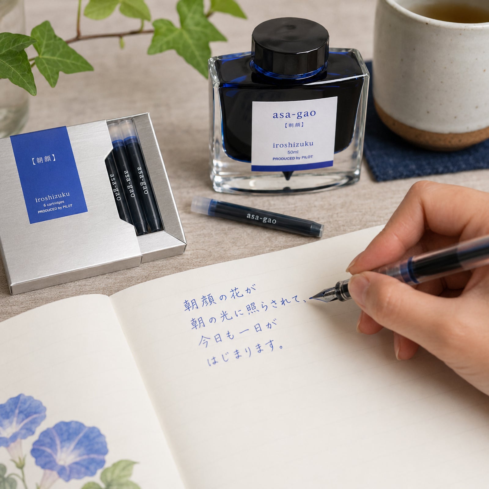 Pilot Iroshizuku Asa gao Morning Glory fountain pen ink cartridges displayed beside the matching 50ml ink bottle while a hand writes with a compatible fountain pen in a notebook. The vibrant blue ink reflects the fresh beauty of the Morning Glory flower and showcases its expressive writing performance.
