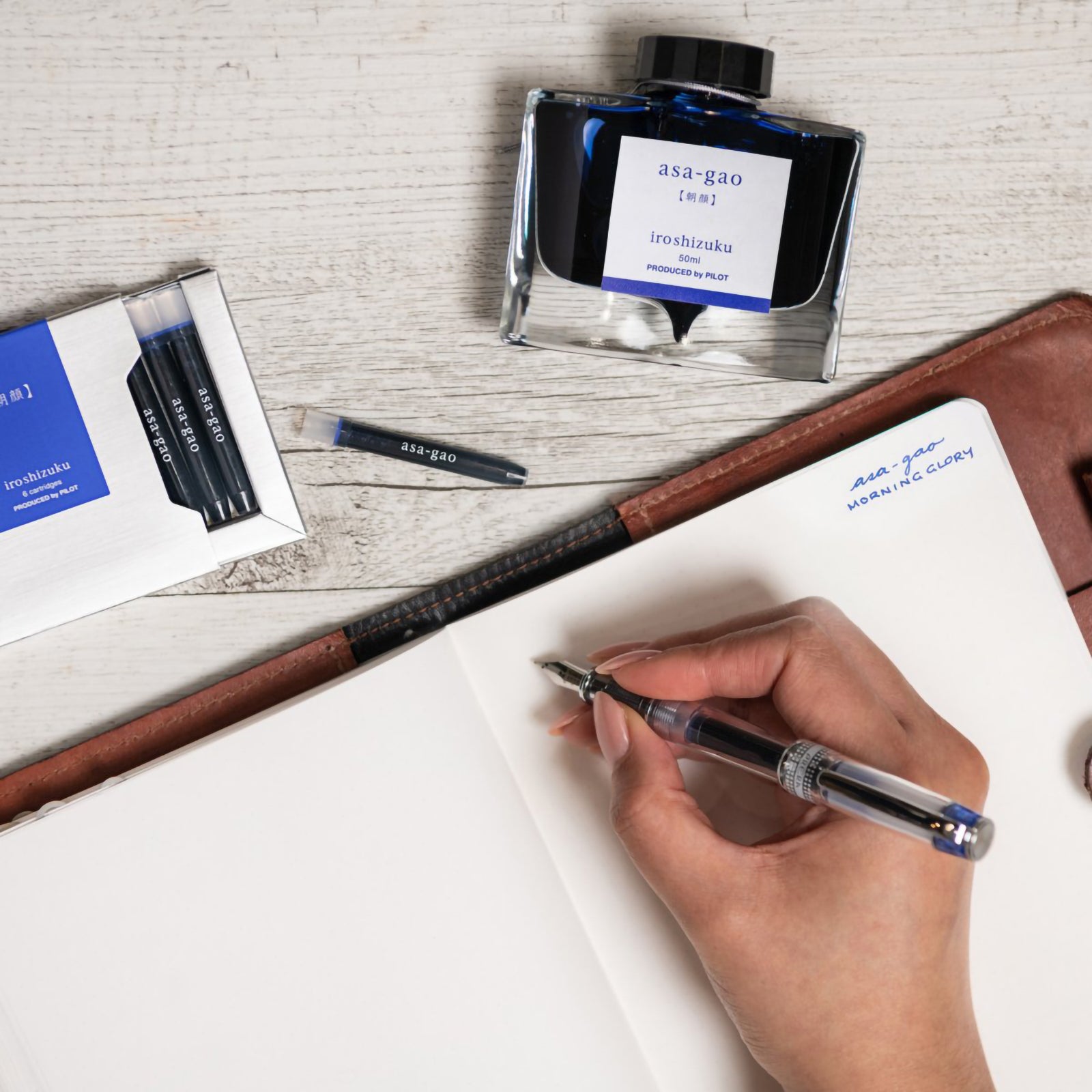 Creative writing workspace featuring the Pilot Iroshizuku Asa gao cartridge box, matching 50ml ink bottle, loose cartridge and compatible fountain pen beside an open notebook. The arrangement demonstrates the premium writing experience inspired by the Morning Glory flower.