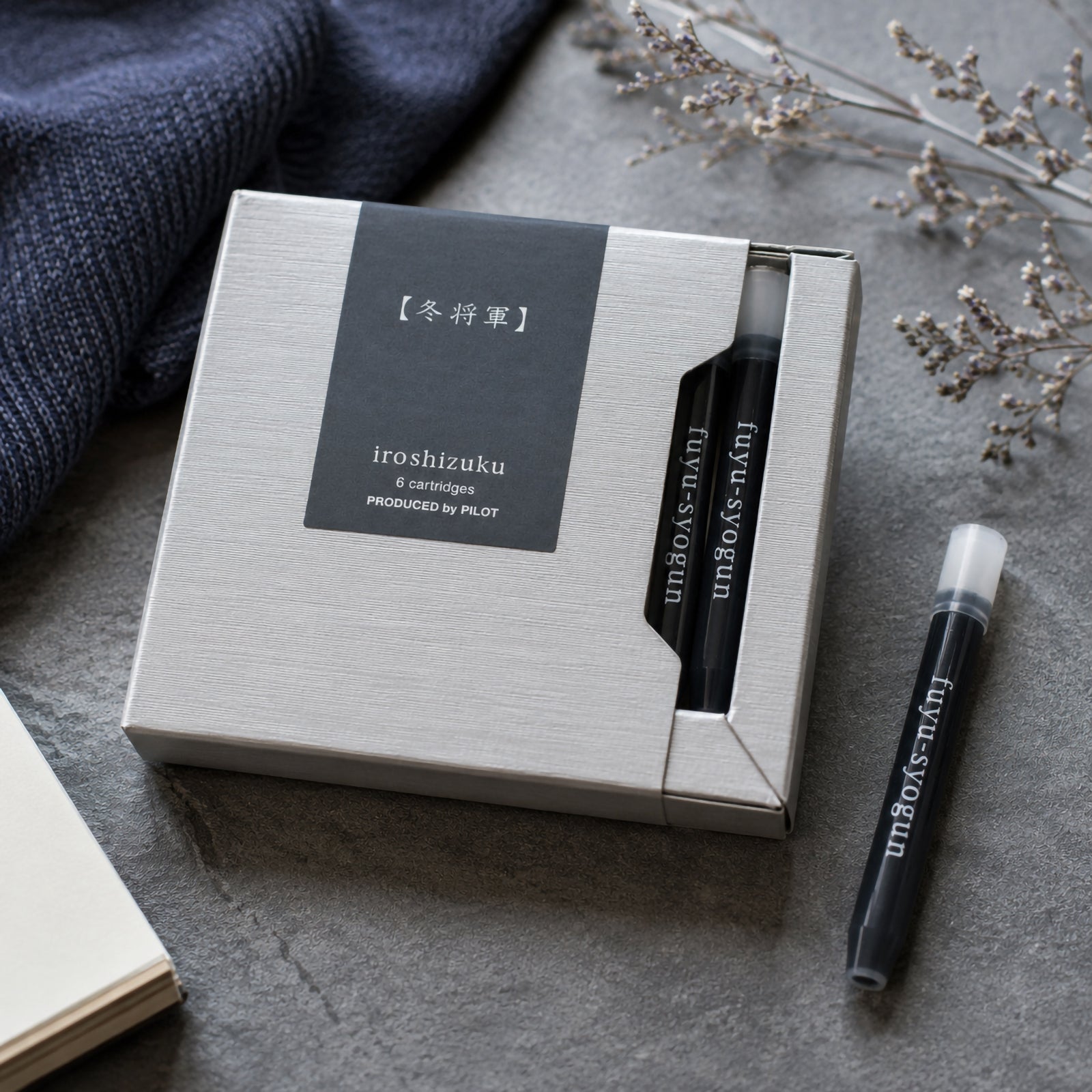 Premium lifestyle presentation of the Pilot Iroshizuku Fuyu syogun six cartridge gift box with two visible cartridges and one loose cartridge resting on a stone surface beside soft textiles and dried botanical accents. The composition showcases the refined winter inspired packaging and sophisticated grey ink.