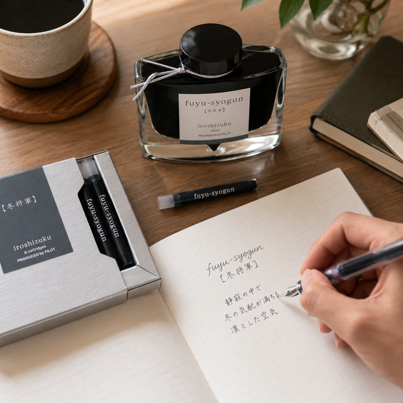 Pilot Iroshizuku Fuyu syogun Old Man Winter fountain pen ink cartridges displayed beside the matching 50ml ink bottle while a hand writes with a compatible fountain pen in an open notebook. The warm timber workspace highlights the elegant cool grey ink inspired by the peaceful atmosphere of a Japanese winter.