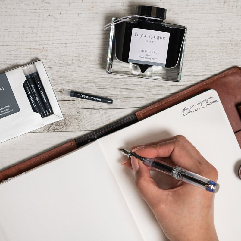 Creative writing workspace featuring the Pilot Iroshizuku Fuyu syogun cartridge box, matching 50ml ink bottle, loose cartridge and compatible fountain pen beside an open notebook. The arrangement demonstrates the premium writing experience inspired by the calm beauty of winter.