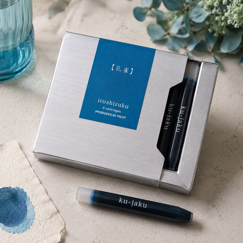 Premium lifestyle display featuring the Pilot Iroshizuku Ku jaku six cartridge presentation box with two visible cartridges and one loose cartridge beside watercolour paper, blue glassware and fresh eucalyptus. The scene showcases the elegant blue green inspired packaging.