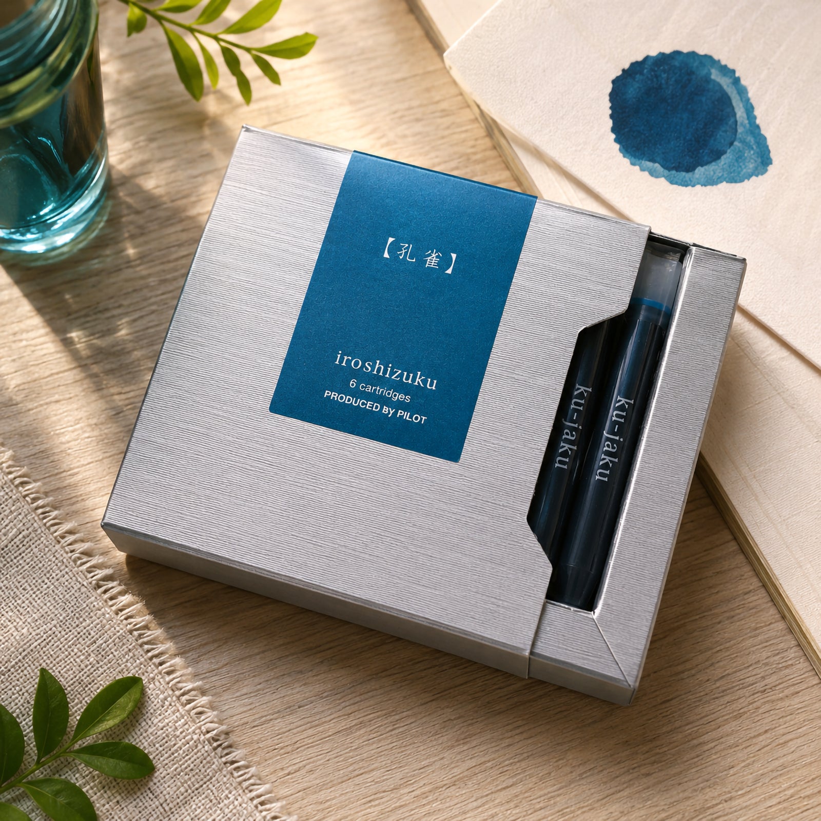 Pilot Iroshizuku Ku jaku cartridge presentation box styled on a timber desktop with botanical accents, blue glassware and colour swatches. The arrangement reflects the vivid peacock inspired ink while highlighting the premium packaging and genuine Pilot cartridges.