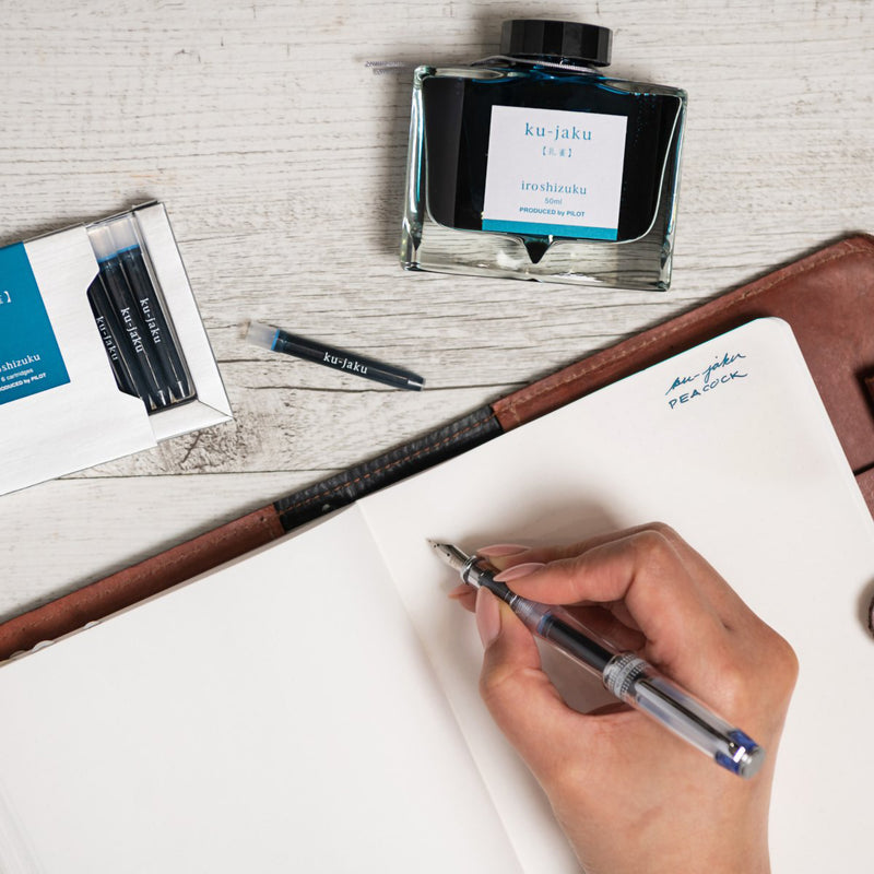 Creative writing workspace showing the Pilot Iroshizuku Ku jaku cartridge box, matching 50ml ink bottle and loose cartridge beside an open journal as a compatible fountain pen is prepared for writing. The scene demonstrates the premium writing experience with rich blue green ink.