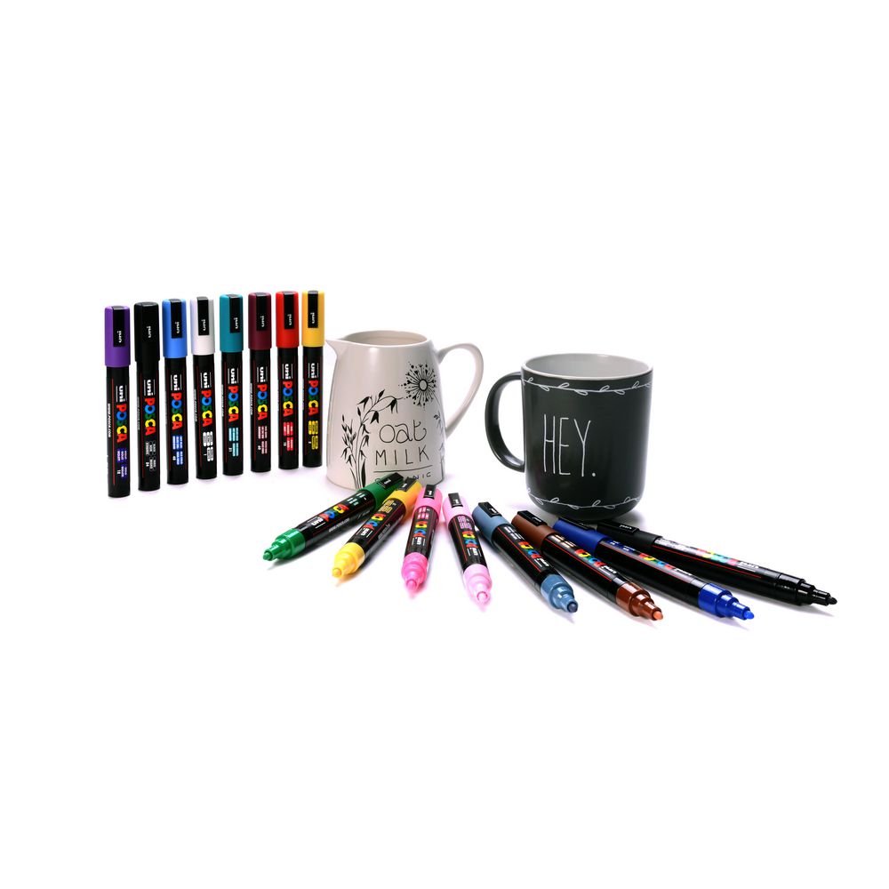 Posca PC-5M markers arranged on a white surface beside a cream jug hand lettered with Oat Milk and a black mug lettered with Hey, with eight capped markers standing upright and eight more lying uncapped.