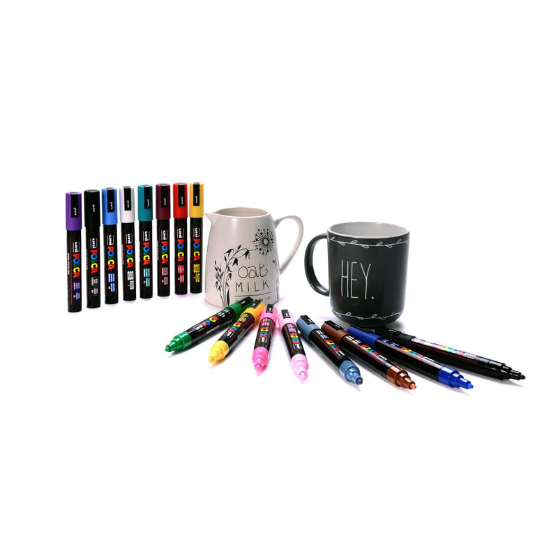 Posca PC-5M markers arranged on a white surface beside a cream jug hand lettered with Oat Milk and a black mug lettered with Hey, with eight capped markers standing upright and eight more lying uncapped.