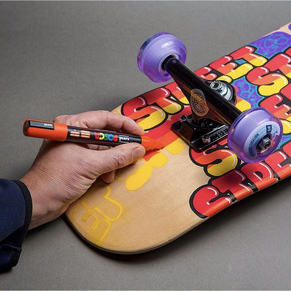A hand uses an orange Posca PC-5M paint marker to fill in yellow bubble lettering on the bare timber underside of a skateboard deck, which already carries red and yellow graffiti style artwork.