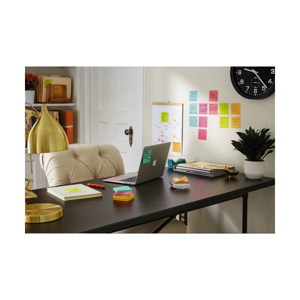 Home office desk by a window with a laptop, a stack of coloured square notes and many notes on the wall behind